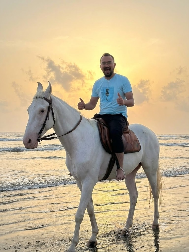 Tanmay riding a white horse on the beach at sunset, both thumbs up